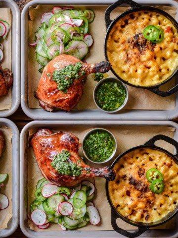 Two trays each with a smoked duck leg, served with a Charred Scallion Chimichurri, mac & cheese, and a cucumber salad.