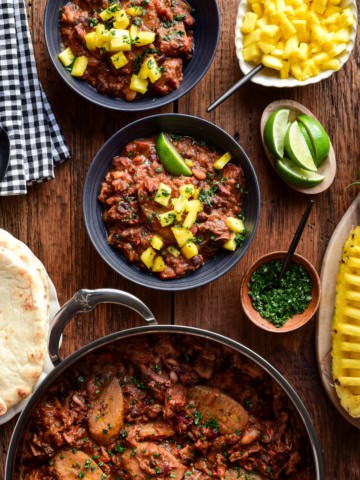 Two bowls and a pot of Caribbean Baked Beans with Jerk-Spiced Pork Shoulder. Served with pineapple, naan and lime wedges.