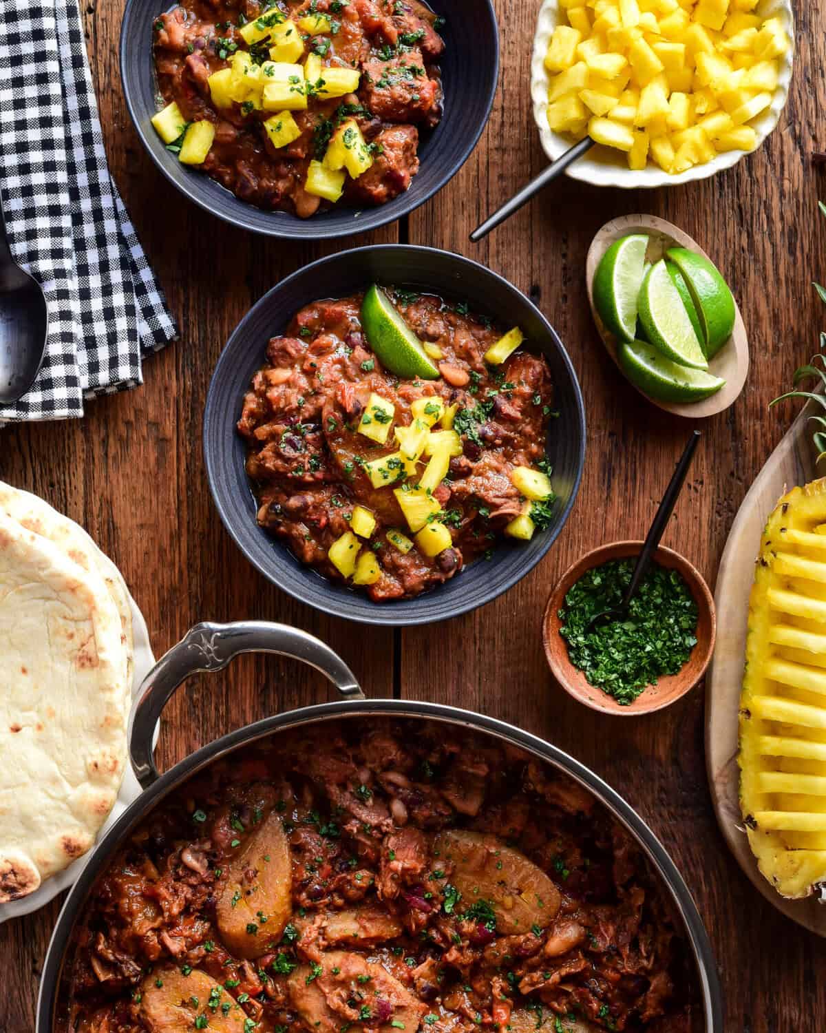Two bowls and a pot of Caribbean Baked Beans with Jerk-Spiced Pork Shoulder. Served with pineapple, naan and lime wedges. 