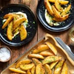 A tray of Greek-Style Lemon Potato Wedges served on plates with whipped feta, chili crisp drizzle, salt and parsley.