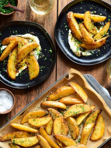 A tray of Greek-Style Lemon Potato Wedges served on plates with whipped feta, chili crisp drizzle, salt and parsley.