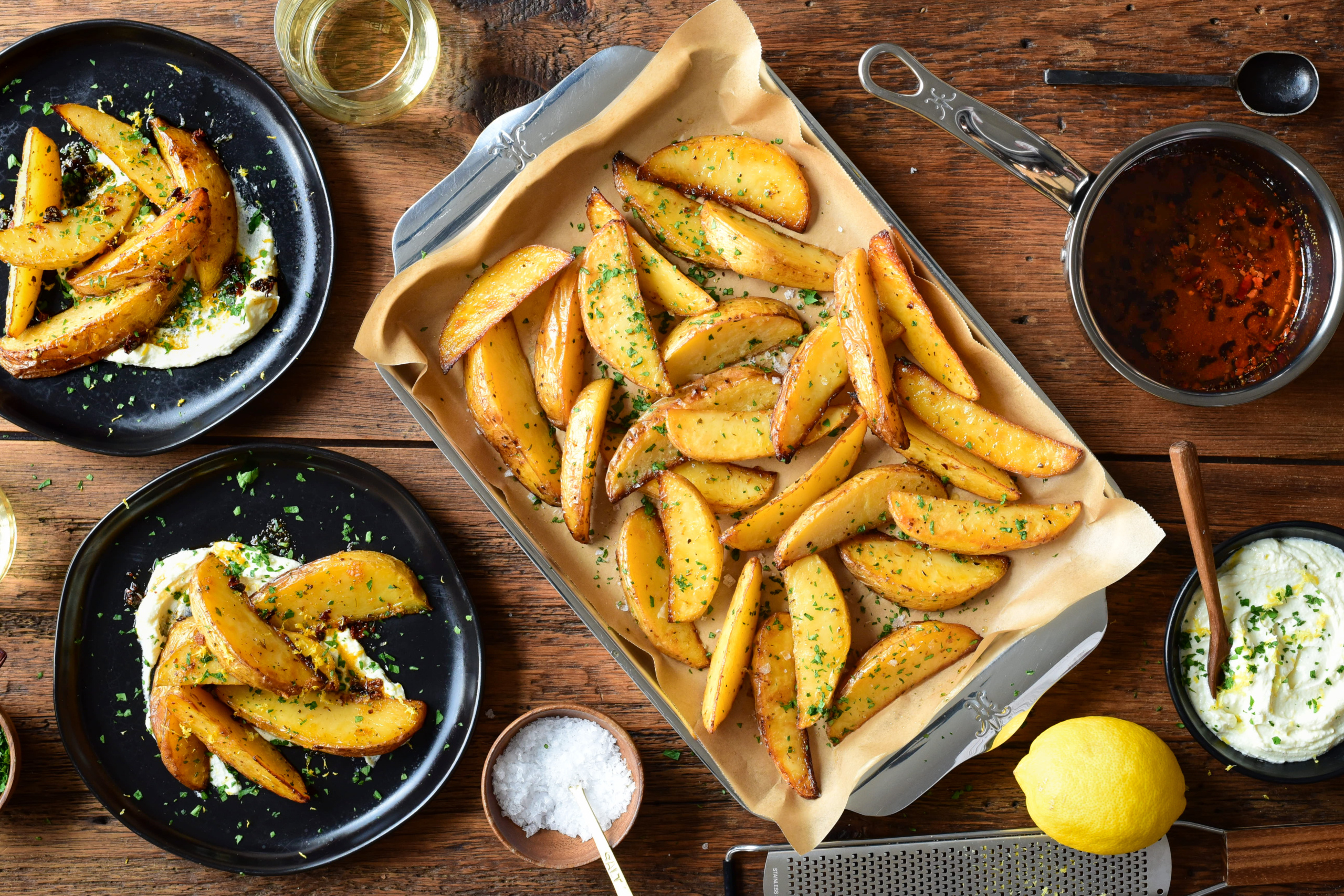 A sheet pan of lemon potato wedges and two portions on a plate served over whipped feta and drizzled with a chili crisp drizzle.