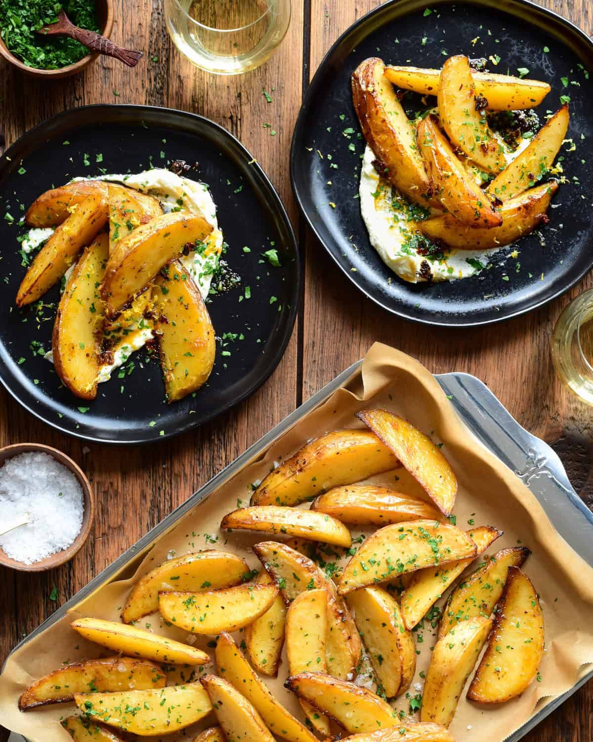 A tray of Greek-Style Lemon Potato Wedges served on plates with whipped feta, chili crisp drizzle, salt and parsley.