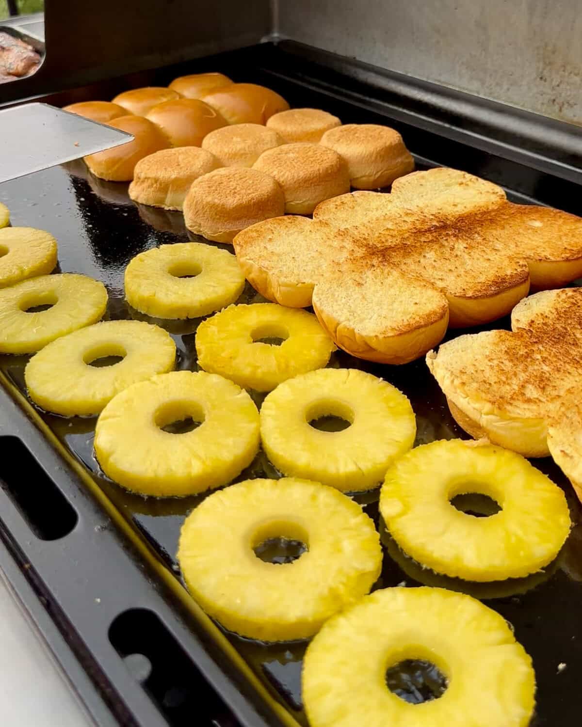 A griddle with circles of pineapple slice and slider buns getting toasted.