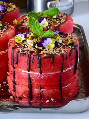 A close-up of mini watermelon cakes with coconut whipped cream, dark chocolate drizzle and topped with pistachios, toasted coconut, mint and edible flowers.