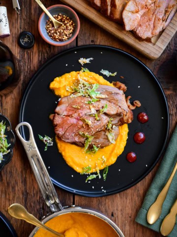 A plate with slices of gochujang honey glazed lamb on a bed of carrot puree. Surrounded by a pot of puree and more lamb slices.