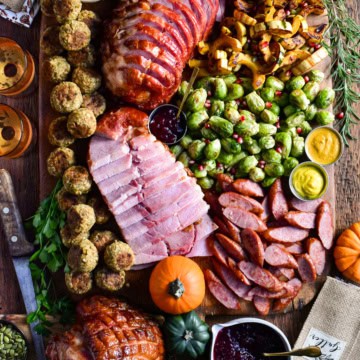 Slices of festive ham and turkey kolbassa are artfully arranged on a board with roasted Brussels sprouts, caramelized delicata squash, stuffing balls, cranberry sauce, and soft dinner rolls. 