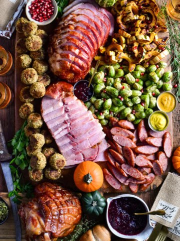 Slices of festive ham and turkey kolbassa are artfully arranged on a board with roasted Brussels sprouts, caramelized delicata squash, stuffing balls, cranberry sauce, and soft dinner rolls.