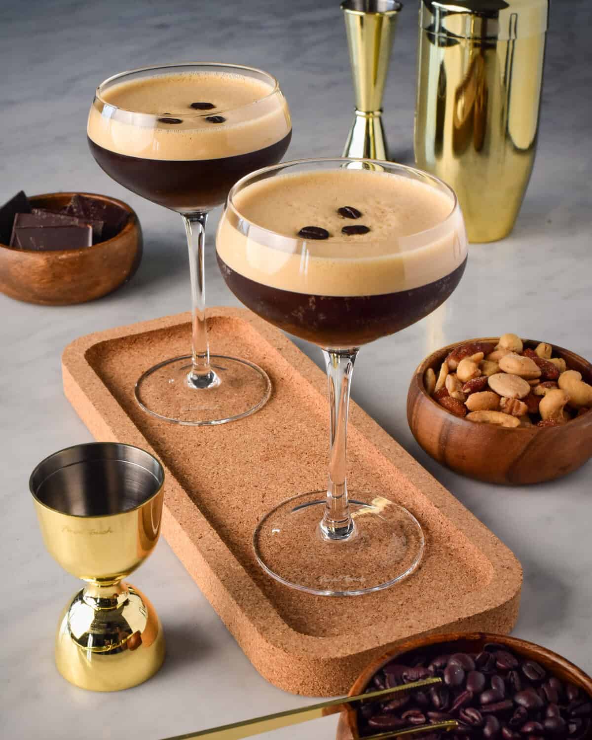 Two espresso martinis on a cork tray, served with nuts and chocolate.