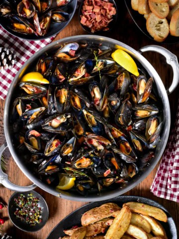 A pot of PEI Mussels in Cider Broth with Bacon and Cranberry Salt. they are served bread, lemon wedges and salt and malt vinegar potato wedges.
