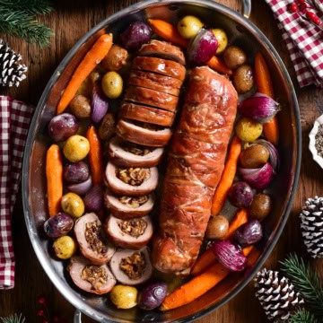 Two Prosciutto Wrapped Pork Tenderloin Stuffed with Figs & Brie (one is sliced) and served in an oval platter with roasted vegetables.