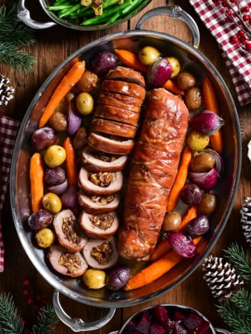Two Prosciutto Wrapped Pork Tenderloin Stuffed with Figs & Brie (one is sliced) and served in an oval platter with roasted vegetables.