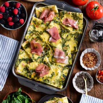 Sheet pan eggs cut into squares, served with prosciutto slices, bagels, arugula, and berries.