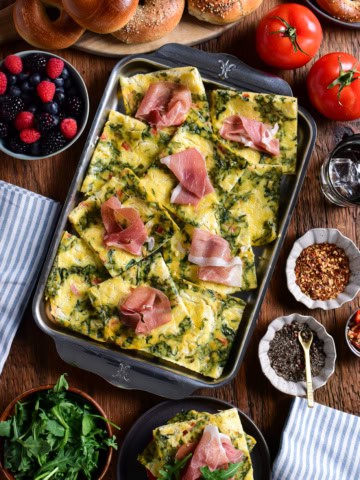 Sheet pan eggs cut into squares, served with prosciutto slices, bagels, arugula, and berries.