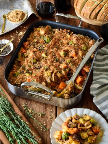 A winter vegetable cassoulet in a roaster with a serving spoon. A portion is served on a plate and accompanied by extra breadcrumb toppings and sourdough bread.