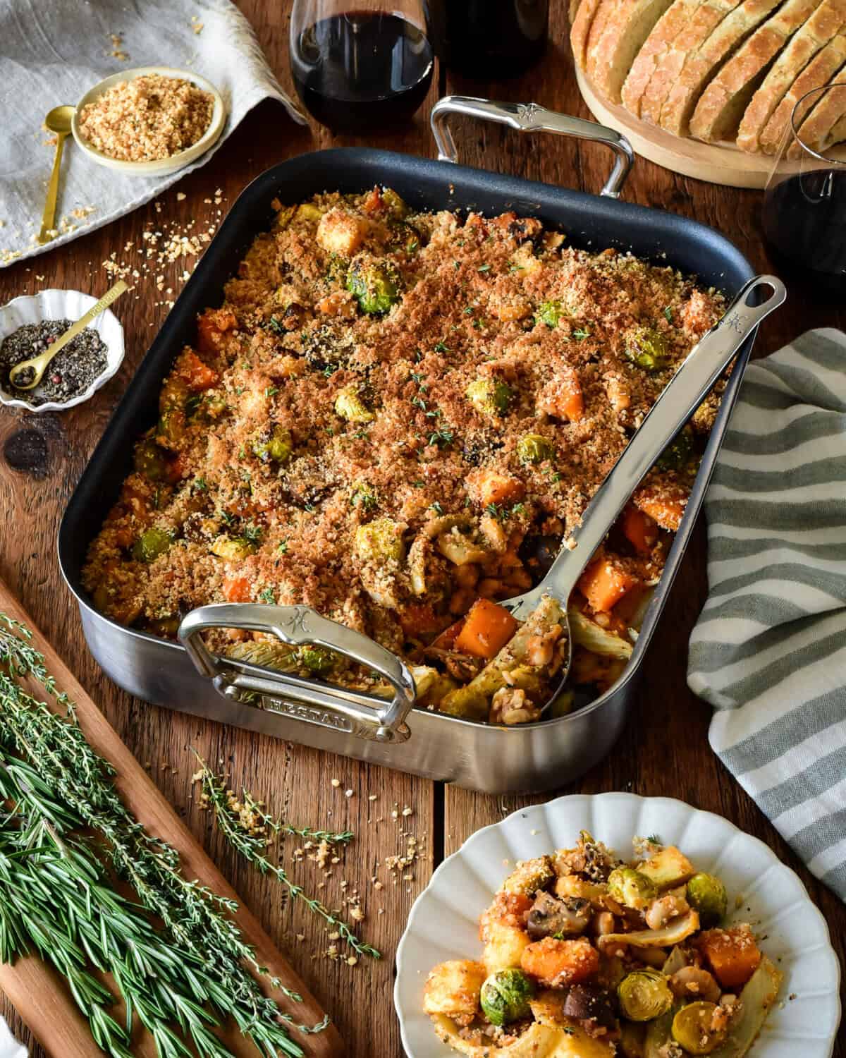a winter vegetable cassoulet in a roaster with a serving spoon. a portion is served on a plate and is served with extra breadcrumb toppings and sourdough bread.
