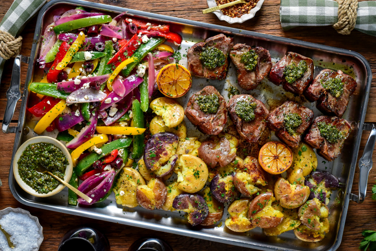 Garlic and anchovy lamb chops with warm salsa verde, served on a stainless-steel griddle with smashed baby potatoes, griddle peppers, and onions.