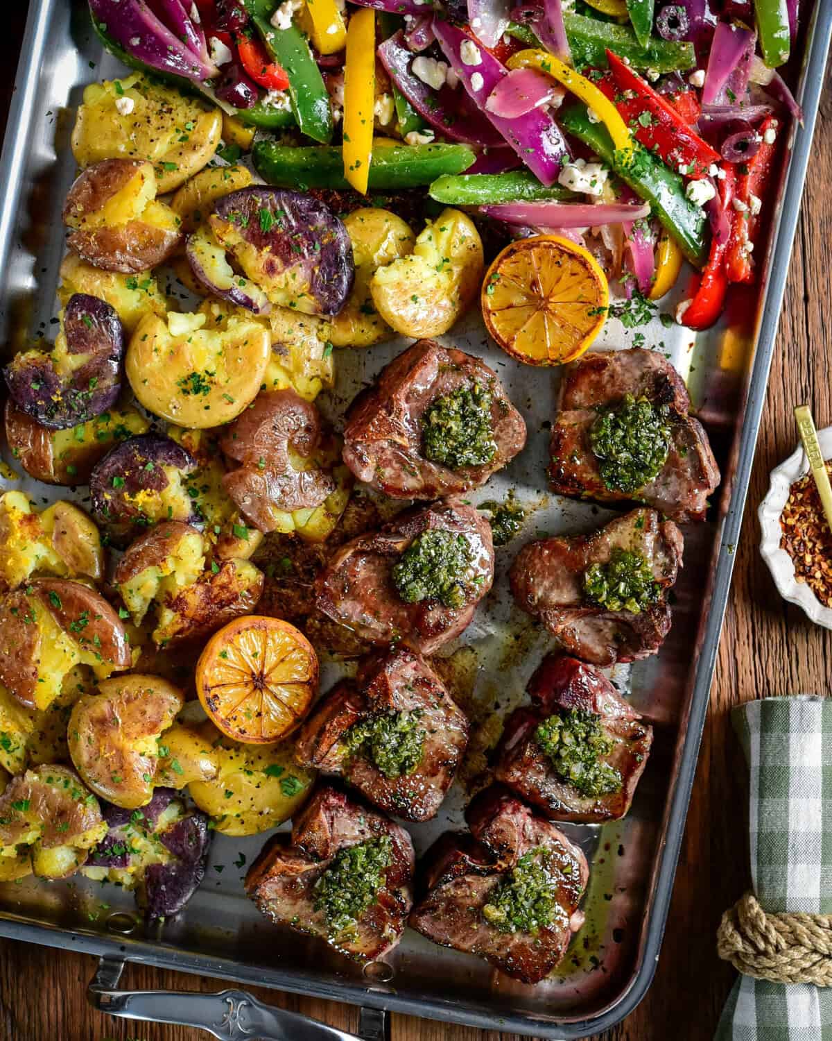 Garlic and anchovy lamb chops with warm salsa verde, served on a stainless-steel griddle with smashed baby potatoes, griddle peppers, and onions.