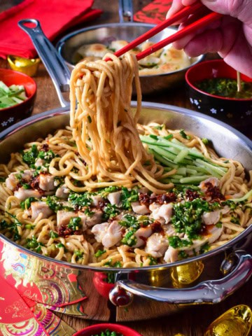A pan of longevity yi mein noodles with ginger chive chicken, getting pulled up with red chopsticks.