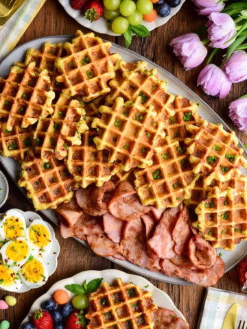 A platter with cornmeal waffles made with ham and cheddar and served with extra old-fashioned ham slices, hard-boiled eggs, fruit, and maple syrup.