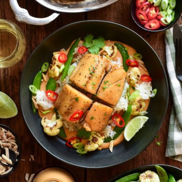 Baked black cod served in a bowl with Thai peanut curry sauce, crispy rice, roasted cauliflower, snap peas, red chilli peppers and a lemon wedge.