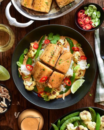 Baked black cod served in a bowl with Thai peanut curry sauce, crispy rice, roasted cauliflower, snap peas, red chilli peppers and a lemon wedge.