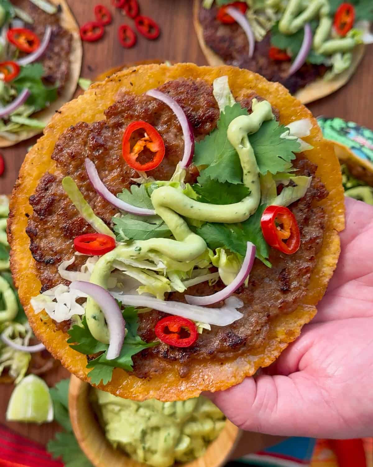 A close-up of crispy pork smash tacos with avocado lime cream, cilantro, onions, lettuce and chiles.
