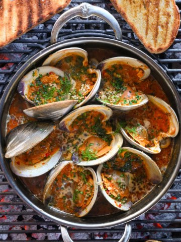 A pot of grilled clams casino in a Calabrian chili sauce over a BBQ with some grilled toast.