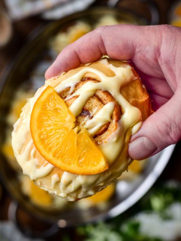 A close-up of a cinnamon roll with orange cream cheese glaze and a piece of candied orange.