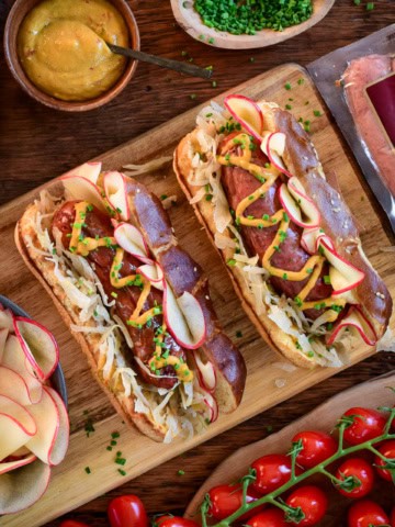 Two charcoal grilled bratwurst with pickled apples, sauerkraut, and mustard on pretzel buns.