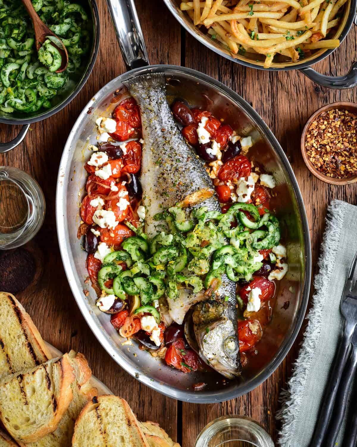A whole roasted branzino in a fish pan with tomatoes, feta and olives. Served with toasted sourdough, a cucumber slaw and shoestring fries.