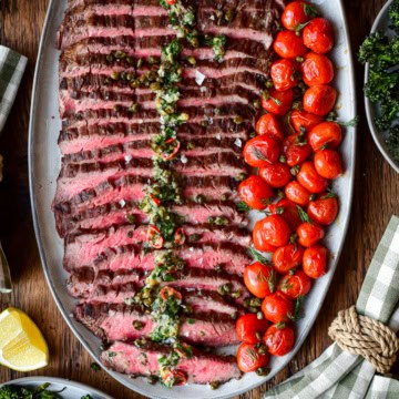 A platter of sliced, grilled, medium-rare flank steak with roasted garlic butter and blistered tomatoes.