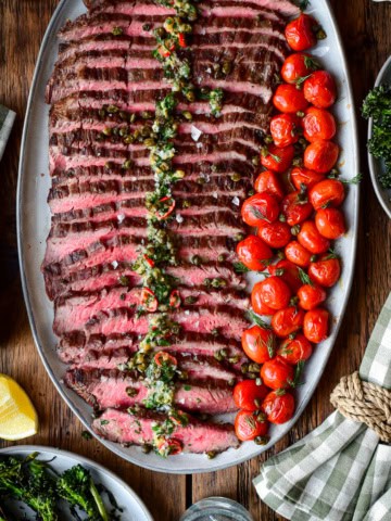 A platter of sliced, grilled, medium-rare flank steak with roasted garlic butter and blistered tomatoes.