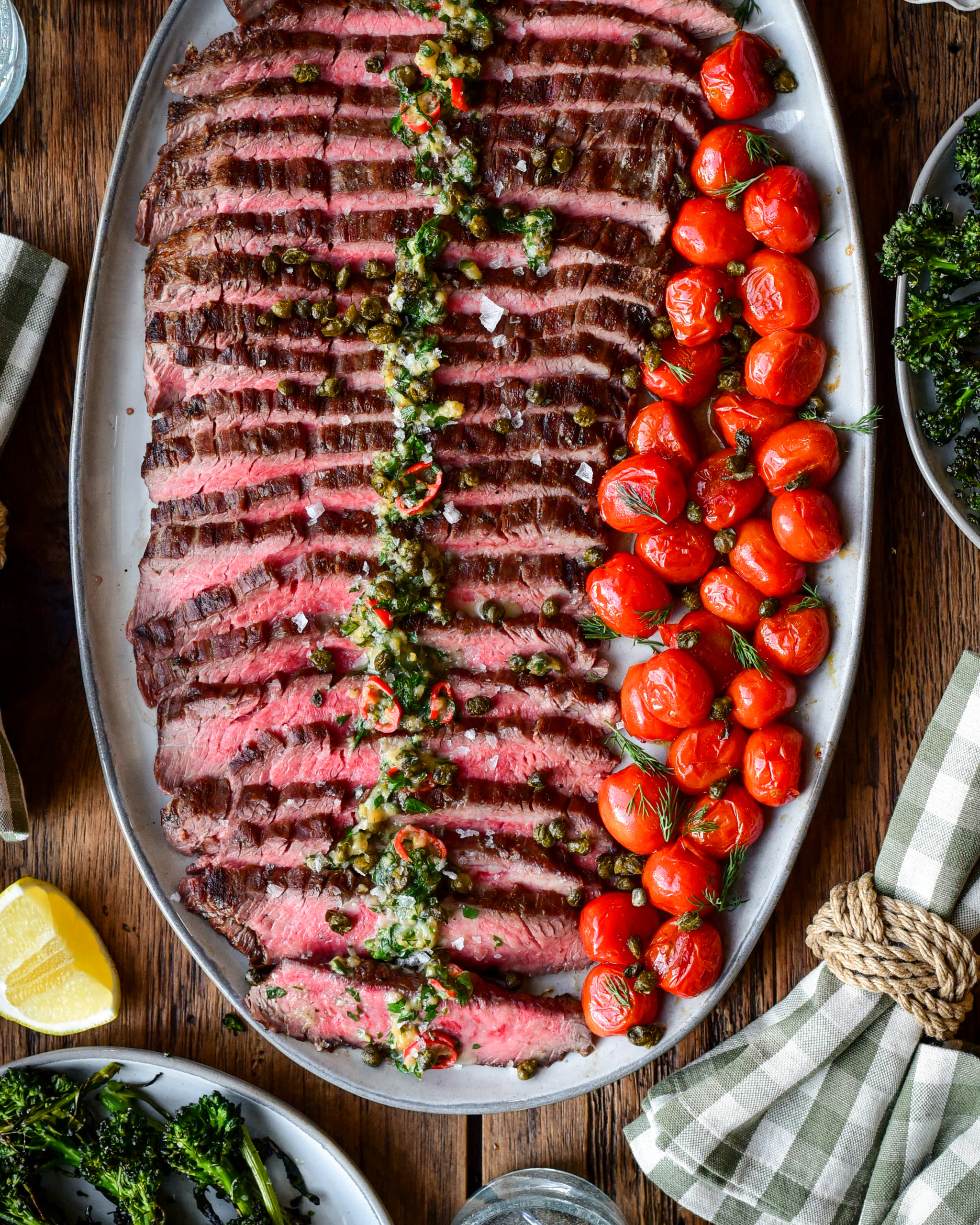 A platter of sliced, grilled, medium-rare flank steak with roasted garlic butter and blistered tomatoes.