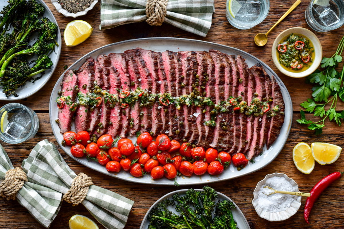 A platter of sliced, grilled, medium-rare flank steak with roasted garlic butter and blistered tomatoes. Served with grilled broccolini and lemon wedges.