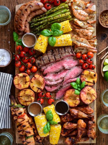 Colorful grilled meats, vegetables, and sides on a wooden board for a BBQ charcuterie platter.
