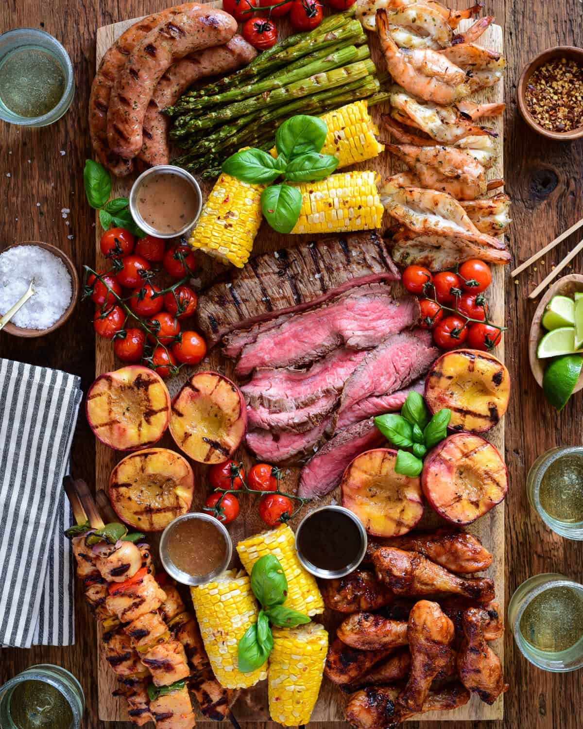 Colorful grilled meats, vegetables, and sides on a wooden board for a BBQ charcuterie platter.