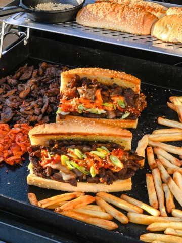 Two bulgogi cheesesteaks on the griddle with fries and extra sandwich ingredients.