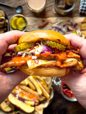 Close-up of a Nashville hot honey veal cutlet sandwich with pickles, coleslaw, and sauce in a bun, held by hands over a rustic wooden table with sides and drinks.