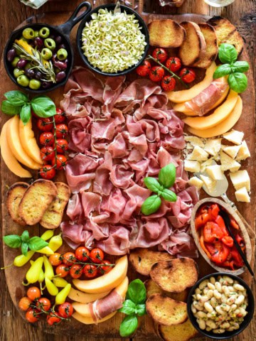 A Prosciutto di Parma Antipasto Butter Board with whipped butter, marinated white beans, melon, marinated olives, tomatoes, grilled bread, parmesan cheese, pickled peppers and fresh basil.