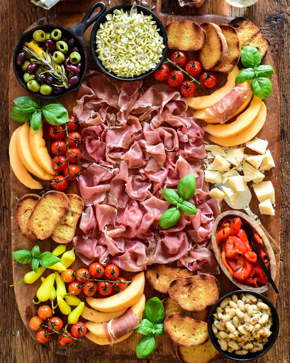 A Prosciutto di Parma Antipasto Butter Board with whipped butter, marinated white beans, melon, marinated olives, tomatoes, grilled bread, parmesan cheese, pickled peppers and fresh basil.