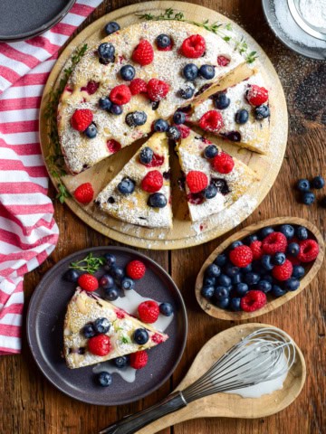 A Lemon Blueberry and Raspberry Cake with blueberries and lemon drizzle on a round serving plate on a rustic wooden table with a red and white striped tea towel. A slice is served with extra berries and glaze, with extra berries on the side.