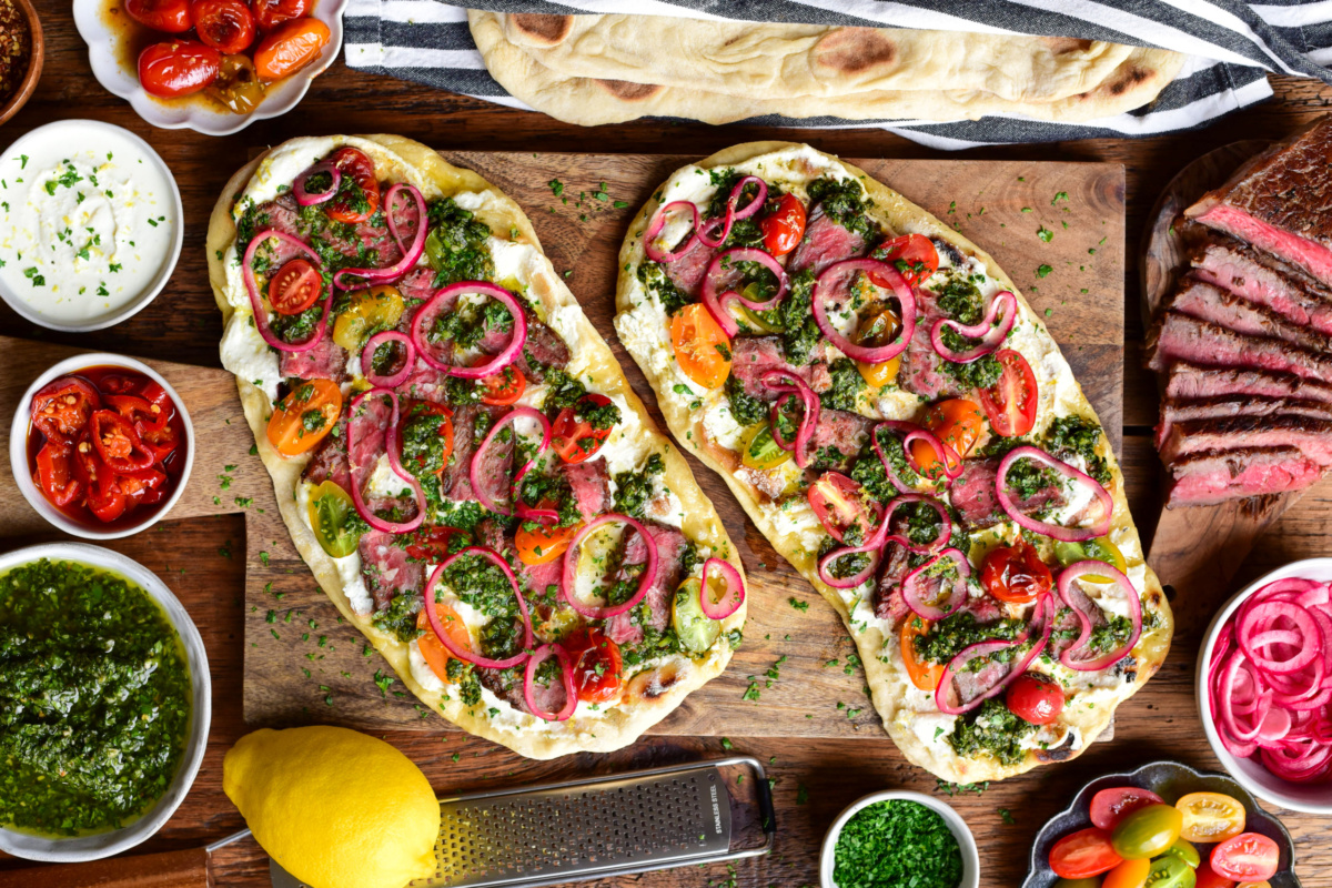 Two chimichurri steak flatbreads with whipped feta, marinated onions and blistered tomatoes on a wooden board. Extra ingredients are placed around this board.