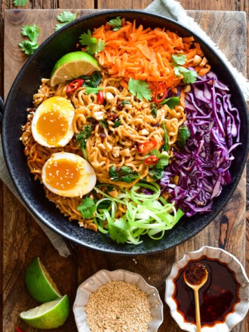 A colourful bowl of saucy chili-crunch almond butter ramen noodles in a large bowl with red cabbage, grated carrots and soft-boiled eggs. Extra chili crunch, lime wedges and toasted sesame seeds are served on the side.