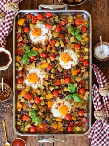 A stainless steel griddle with an Italian sausage and pepper breakfast hash with four fried eggs.