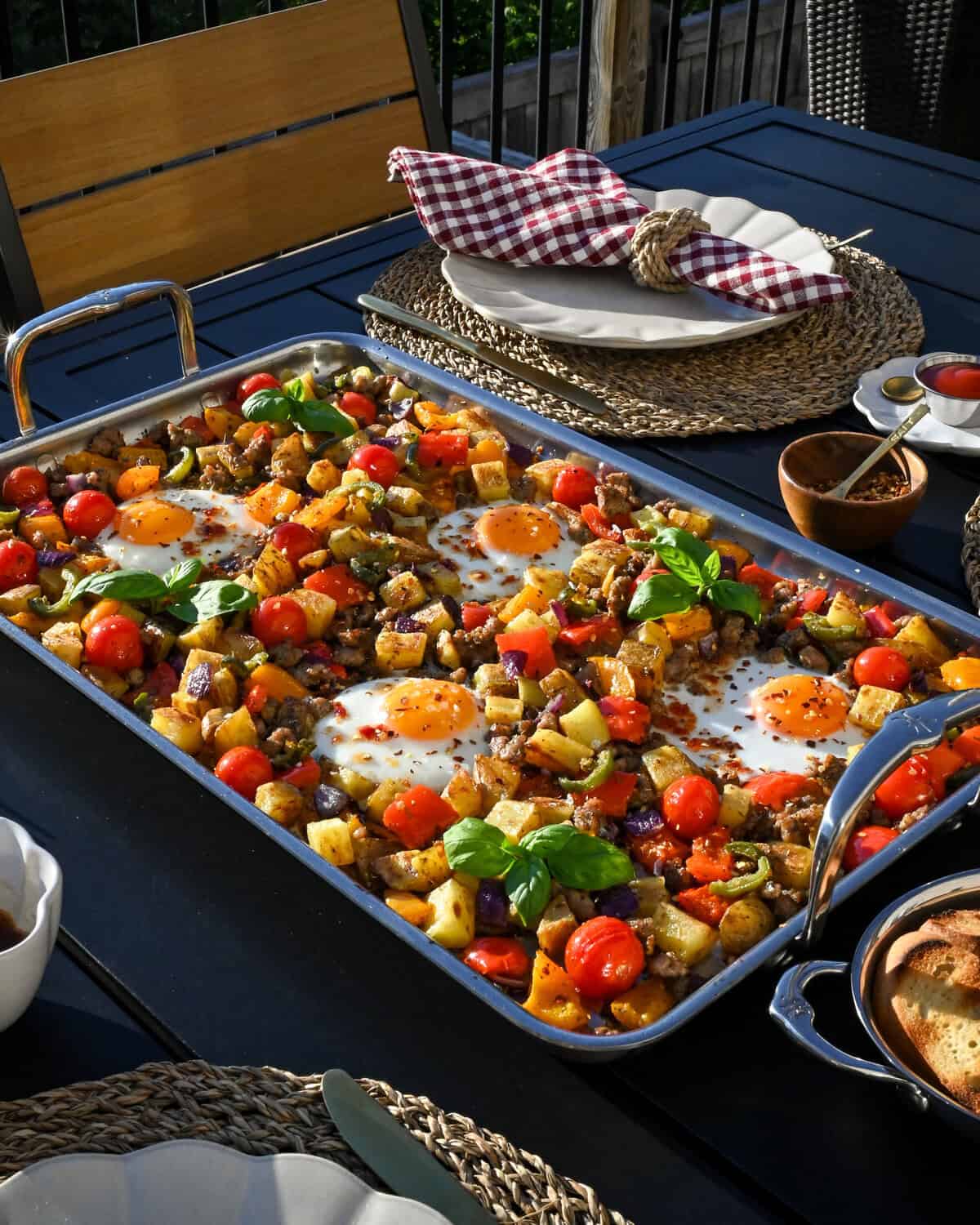 A stainless steel griddle with an Italian sausage and pepper breakfast hash with four fried eggs. Served outdoors with a table set.