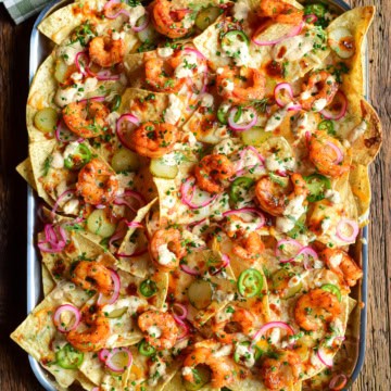 A tray of Nashville Hot Honey Shrimp Nachos with Pickle Crema. Layered with pickled onions, jalapenos, dill pickle slices and herbs.
